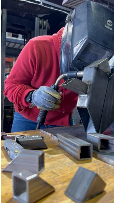 A welder in a red hoodie and welding helmet running a weld along a steel bracket at the shop bench, cut lengths of square tubing laid out in front.