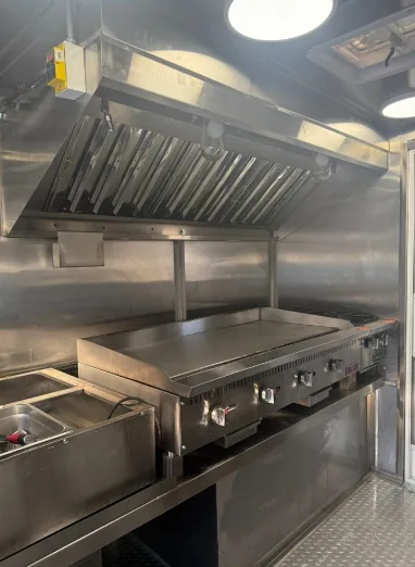 A stainless steel extraction hood over a flat-top griddle and burner range inside a mobile kitchen, with a steam table beside them.