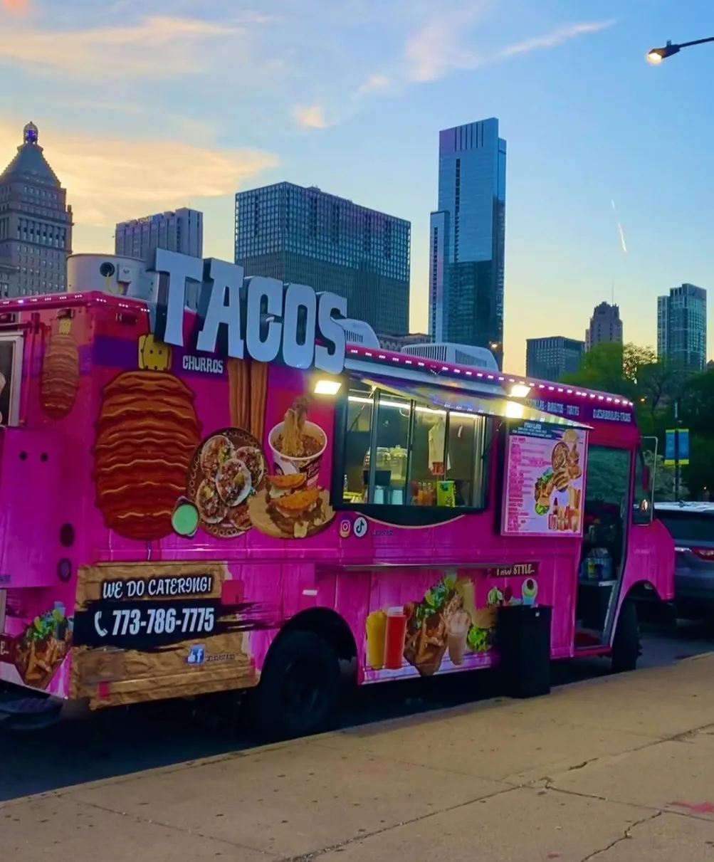 A pink taco truck with three-dimensional lettering on its roof, serving hatch open at dusk with a city skyline behind it
