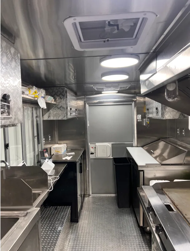The finished stainless steel interior of a mobile kitchen — prep counters and sink down one side, cooking line down the other, diamond-plate floor running back to the rear door.
