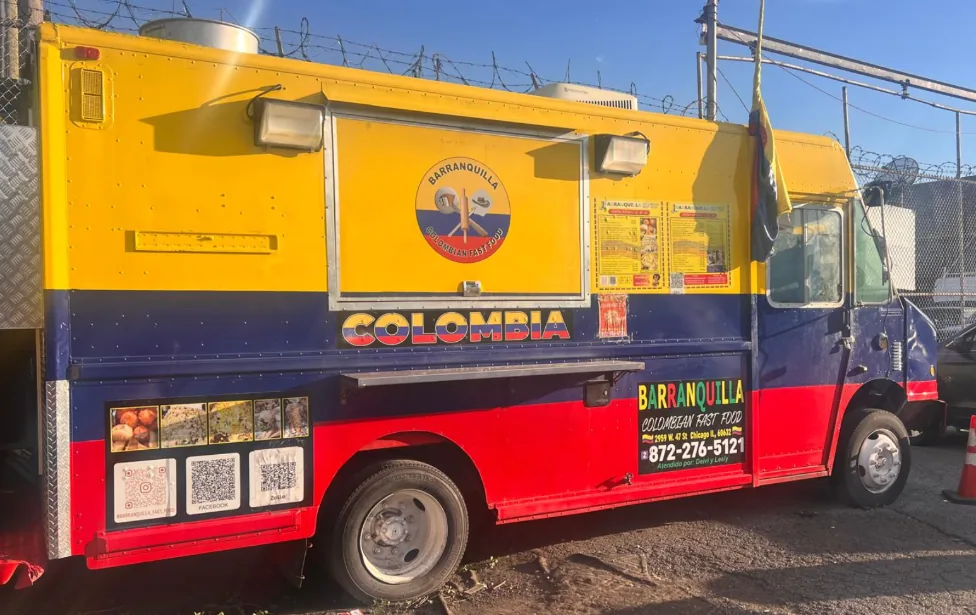 A finished food truck in a full-color yellow, blue and red wrap, parked in a lot on a sunny day with its serving window propped open and menu boards mounted beside it.