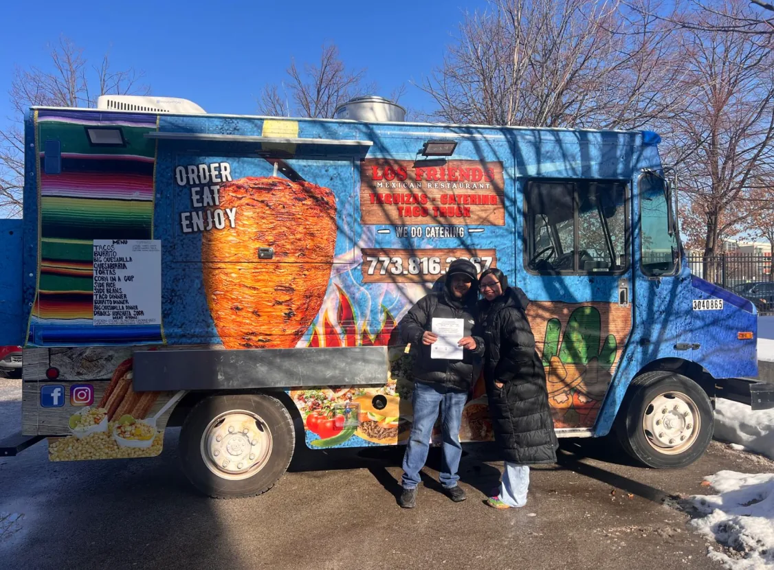 Two owners standing in front of their finished taco truck, wrapped in a full-color al pastor design, holding a certificate up for the camera on a bright winter morning.