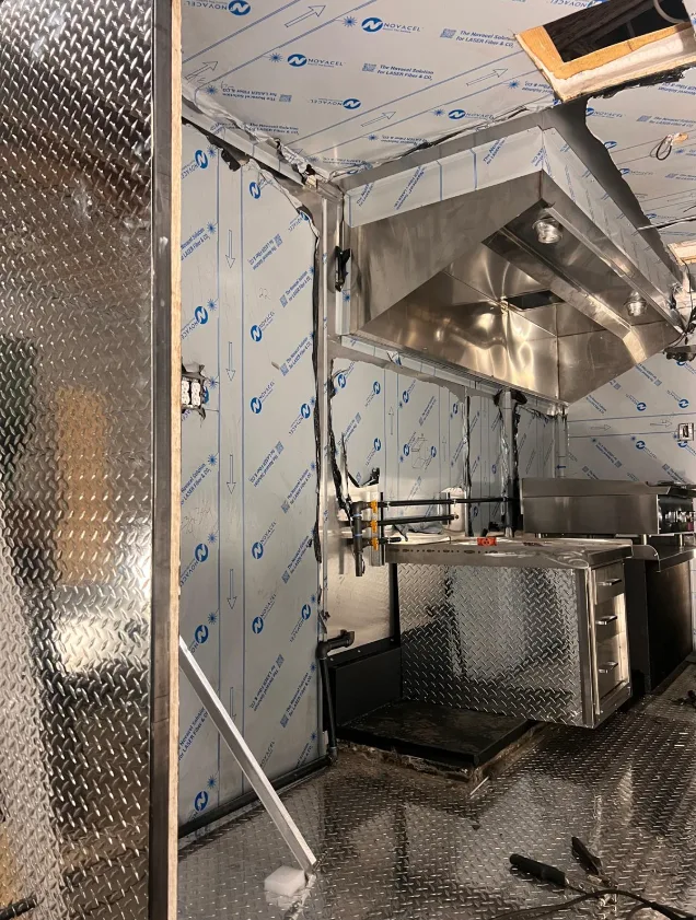 A stainless steel extraction hood and prep counter installed inside a unit part-way through its build, the wall panels still under blue protective film and the gas line run to the cooking position.