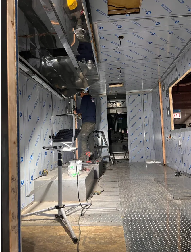 A worker on a step inside a unit part-way through its fit-out, fixing the stainless ceiling panel beside a newly hung extraction hood, the walls lined with insulation board over timber framing and a work light standing on a tripod.