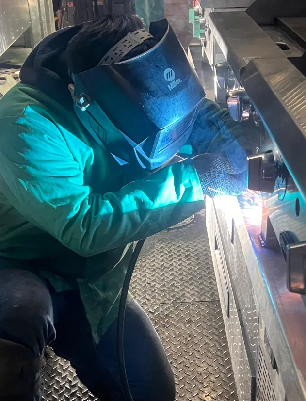 A welder in a green flame-resistant jacket kneeling on a diamond-plate floor inside a unit, welding a stainless steel counter frame as sparks fly.