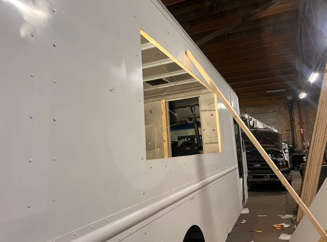 A service window cut into the side of a white step van in the shop, the opening framed out in fresh timber, with the workshop's brick wall and timber ceiling behind it.