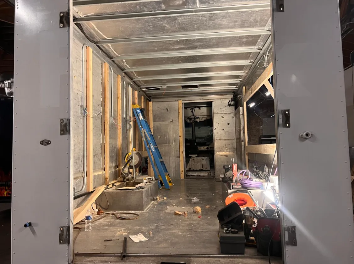 The bare shell of a box truck on the shop floor with its rear doors open, stripped back to the frame and framed out in new timber studs down both walls, a ladder standing inside.