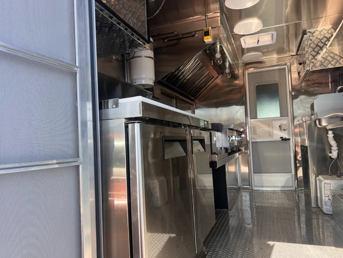 The finished stainless steel interior of a mobile kitchen — a reach-in refrigerator and prep counter down one side, the extraction hood over the cooking line at the far end, a hand sink and water heater opposite, and a diamond-plate floor throughout.