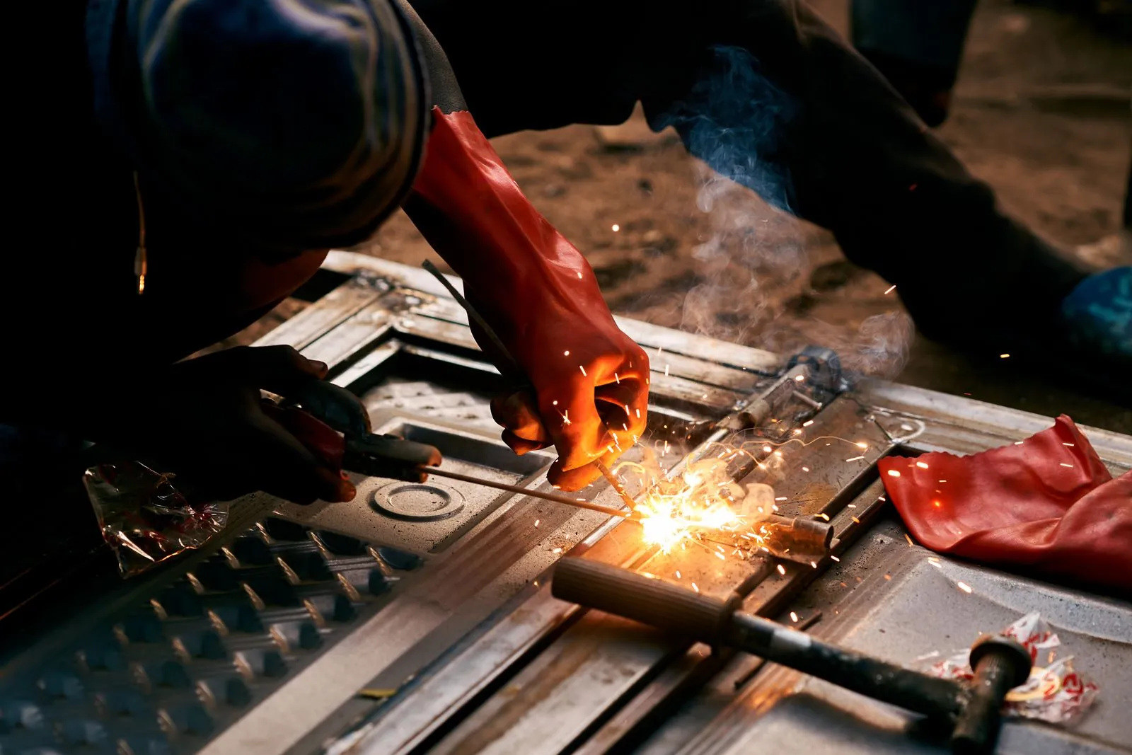 A worker in heavy protective gloves welding a steel part on a bench, sparks flying